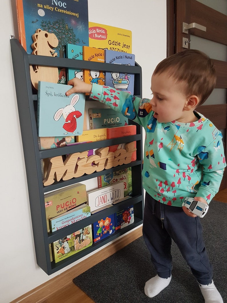 May include: A dark gray wooden bookshelf with multiple shelves filled with children's books. The bookshelf is mounted to a white wall. A young child is standing in front of the bookshelf, reaching for a book. The bookshelf has the name "Micha" spelled out in wooden letters.