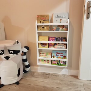 May include: White wall-mounted bookshelf with five shelves holding a variety of children's books. The books have colorful covers with illustrations and titles in Polish. A white door is visible to the right of the bookshelf.