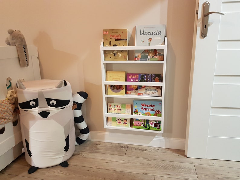 May include: White wall-mounted bookshelf with five shelves holding a variety of children's books. The books have colorful covers with illustrations and titles in Polish. A white door is visible to the right of the bookshelf.