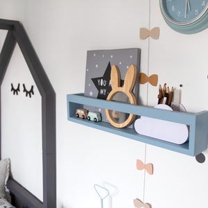 May include: A light blue wall shelf with decorative items, including a wooden bunny, star-shaped artwork, and toy cars. A clock and bow-shaped decorations are also visible. The shelf is mounted on a white wall, creating a playful nursery theme.