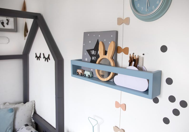May include: A light blue wall shelf with decorative items, including a wooden bunny, star-shaped artwork, and toy cars. A clock and bow-shaped decorations are also visible. The shelf is mounted on a white wall, creating a playful nursery theme.