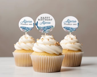 May include: Three cupcakes with white frosting and gold sprinkles. Each cupcake has a round topper with a watercolor whale design and the text "Alfonso Turned One" or "Happy Birthday Alfonso".