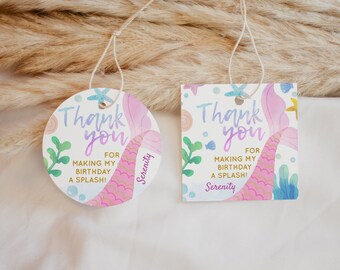 May include: Two round thank you tags with a mermaid tail design. The tags are white with pink, blue, and gold accents. The text on the tags reads "Thank you for making my birthday a splash! Serenity".