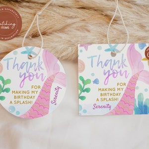 May include: Two round thank you tags with a mermaid tail design. The tags are pink, purple, and blue with gold glitter accents. The text on the tags reads "Thank you for making my birthday a splash!" and "Serenity".