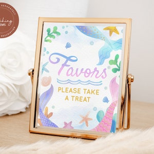 May include: A gold-framed sign with a watercolor mermaid tail design. The sign says "Favors Please Take A Treat" in a pink and blue gradient font.