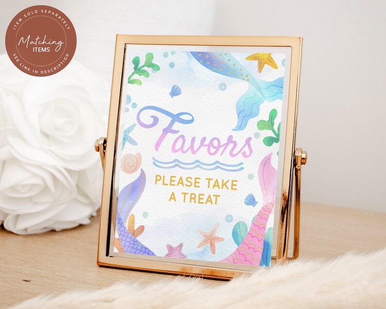 May include: A gold-framed sign with a watercolor mermaid tail design. The sign says "Favors Please Take A Treat" in a pink and blue gradient font.
