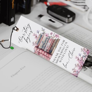Op de afbeelding: Een witte bladwijzer met een stapel vintage boeken, roze bloemen en de tekst "Our story begins" en "Thank you for showering us with love! love, Claire".