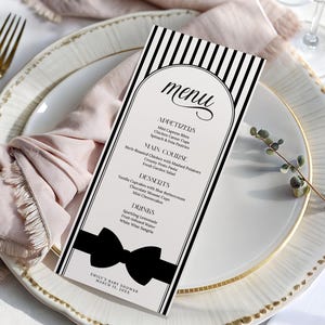 May include: A black and white menu card with a bow design, listing appetizers, main courses, desserts, and drinks. The menu is on a white plate with a gold rim, next to a pink napkin and a gold fork.