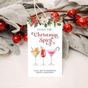 Op de afbeelding: Witte cadeaukaart met een rode en groene bloemenrand. De tekst luidt "Geniet van de kerstsfeer" in rode cursieve letters. Onder de tekst staan drie aquarel-illustraties van cocktails. Onderaan de kaart staat "Love, Ray & Georgina Smith-Anderson".