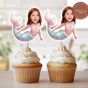 May include: Two cupcakes topped with custom mermaid cake toppers featuring a child's face on a mermaid body with a pink ruffled top and a shimmering iridescent tail. A circular brown badge in the corner contains the text Matching Items.