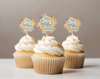 May include: Three cupcakes with white frosting and gold sprinkles topped with round paper cupcake toppers that say "Here comes the sun" in a retro font. The toppers are blue, yellow, and orange.