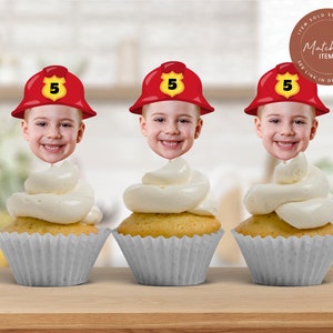 May include: Three red and white firefighter hat cupcake toppers with a child's face in each hat. The hats have a yellow number 5 on them. The toppers are on top of white frosted cupcakes in grey cupcake liners.