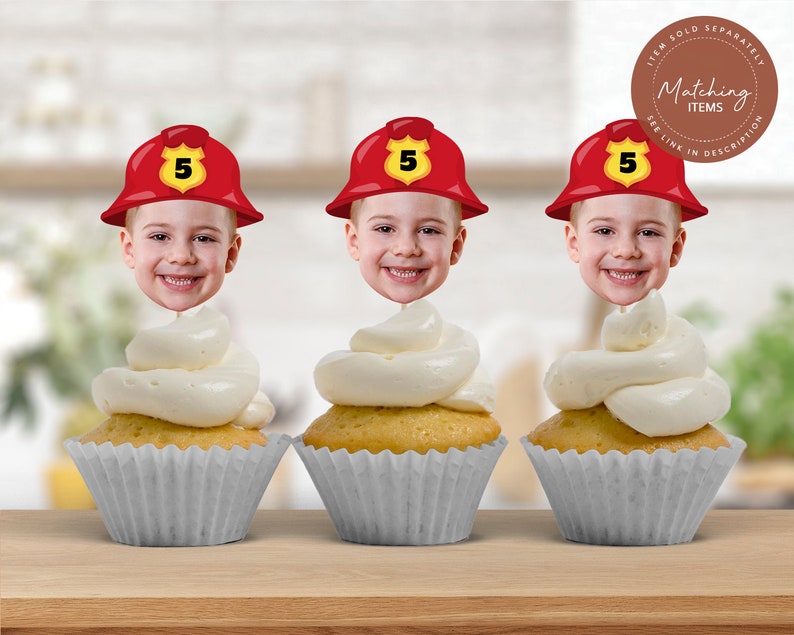 May include: Three red and white firefighter hat cupcake toppers with a child's face in each hat. The hats have a yellow number 5 on them. The toppers are on top of white frosted cupcakes in grey cupcake liners.