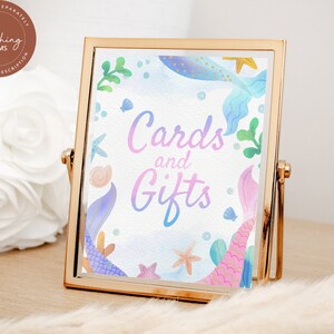 May include: A gold-framed sign with a watercolor background featuring mermaid tails, seashells, and starfish. The sign says "Cards and Gifts" in a pink and blue gradient.