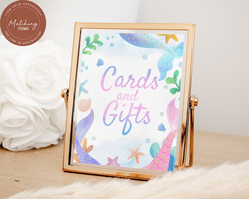 May include: A gold-framed sign with a watercolor background featuring mermaid tails, seashells, and starfish. The sign says "Cards and Gifts" in a pink and blue gradient.