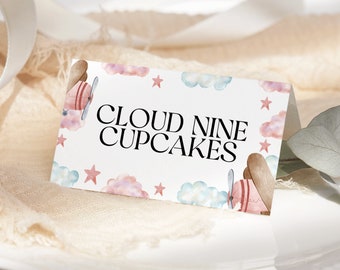 Puede incluir: Una tarjeta plegada con el texto "CLOUD NINE CUPCAKES" en negro. La tarjeta está decorada con ilustraciones de acuarela de aviones, nubes y estrellas en colores pastel. La tarjeta está sobre una superficie blanca.
