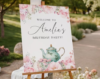 May include: A sign that reads "Welcome to Amelia's Birthday Party!" features a watercolor illustration of a teal teapot, pink flowers, and macarons. The sign is displayed on a wooden easel, with a white tablecloth draped below.