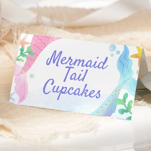May include: A white card with a blue and pink mermaid tail design. The text on the card reads "Mermaid Tail Cupcakes".