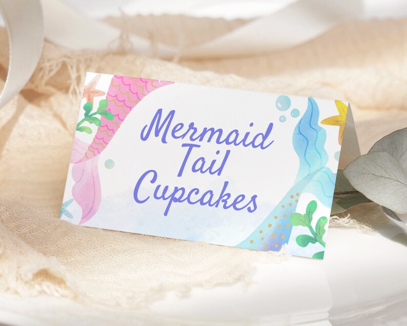May include: A white card with a blue and pink mermaid tail design. The text on the card reads "Mermaid Tail Cupcakes".