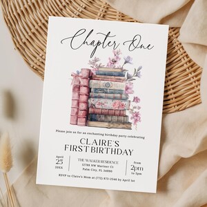 May include: A white rectangular birthday party invitation card featuring the text Chapter One at the top, a watercolor illustration of a stack of vintage books adorned with pink and purple flowers, and event details for Claire's First Birthday.