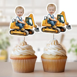 Puede incluir: Dos toppers de cupcake de vehículo de construcción amarillo de juguete con la cara de un niño en cada uno. Los toppers están encima de cupcakes glaseados blancos con chispas naranjas.