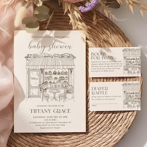 Op de afbeelding: Een uitnodigingsset voor een babyshower met een Parijs thema. De hoofduitnodiging toont een gedetailleerde illustratie van een café met de tekst "baby shower". Bijbehorende kaarten zijn onder meer "Books for Baby" en "Diaper Raffle". De set ligt op een geweven placemat.