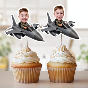 Puede incluir: Dos cupcakes cubiertos con glaseado blanco y chispas doradas. Cada uno tiene un adorno decorativo con una imagen de dibujos animados de un niño en un avión de combate gris. El avión tiene un piloto sonriente.