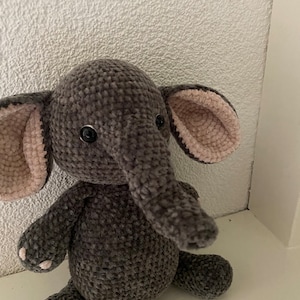 May include: A gray crocheted elephant plush toy with pink ears and black eyes. The elephant is sitting on a white surface.