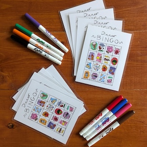 Puede incluir: Un conjunto de cinco tarjetas de bingo en blanco con el título "Dinner Bingo" impreso en cada tarjeta. Las tarjetas están rodeadas de marcadores de colores y una mesa de madera.