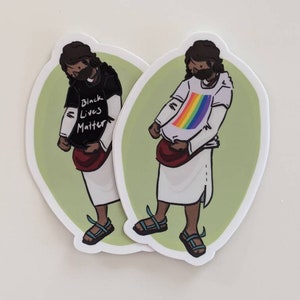 Puede incluir: Dos pegatinas con una ilustraci&oacute;n de dibujos animados de una persona que lleva una t&uacute;nica y sandalias. Una pegatina muestra a la persona con una camiseta negra con el texto "Black Lives Matter" y la otra pegatina muestra a la persona con una camiseta blanca con un arco&iacute;ris.