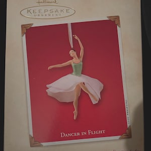 May include: Hallmark Keepsake Ornament box featuring a ballerina in a green leotard and white tutu, mid-flight against a red background. The box has gold-toned corner accents and the text "Dancer in Flight". The box also has the text "Christmas 2003".