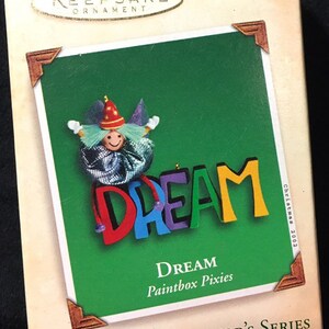 May include: A Christmas ornament featuring a colorful clown holding a word spelled out in large letters that says "DREAM". The ornament is part of a set of two ornaments called "Paintbox Pixies" and is part of the "Miniature Collector's Series".