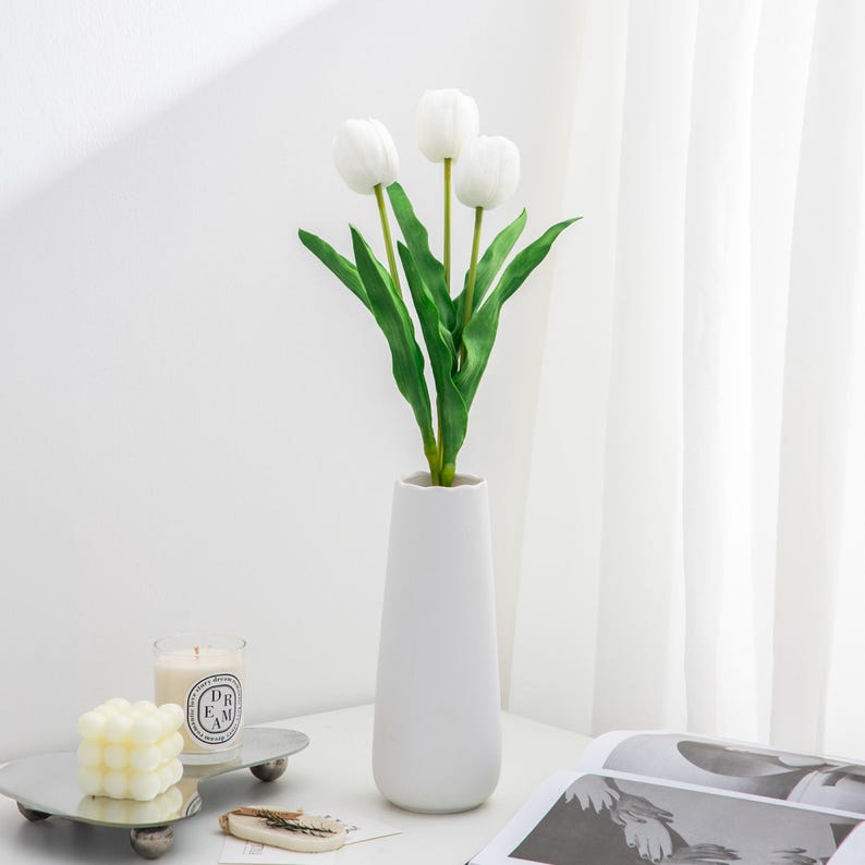 May include: A white ceramic vase with a narrow opening and a wide base, filled with three white artificial tulips with green stems and leaves.