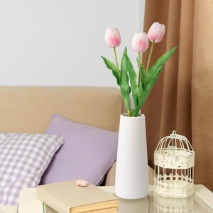 May include: A white ceramic vase containing a bouquet of pink tulips. The vase is on a wooden table with a book and a birdcage.