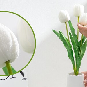 May include: A bouquet of white artificial tulips with green stems in a white vase. The tulips are arranged in a fan shape and the vase is on a white surface.