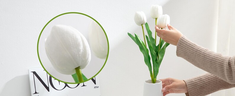 May include: A bouquet of white artificial tulips with green stems in a white vase. The tulips are arranged in a fan shape and the vase is on a white surface.