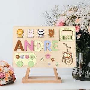 May include: Wooden puzzle board with the name "ANDRE" spelled out in colorful letters. The board features animal shapes, a bus, a helicopter, and a zoo. The puzzle is displayed on a small wooden easel.