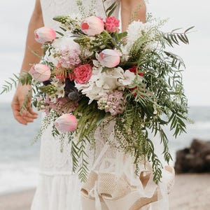 May include: A bridal bouquet featuring pink and white flowers, including tulips, hydrangeas, and greenery. The bouquet is held by a person wearing a white dress.