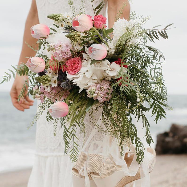 May include: A bridal bouquet featuring pink and white flowers, including tulips, hydrangeas, and greenery. The bouquet is held by a person wearing a white dress.