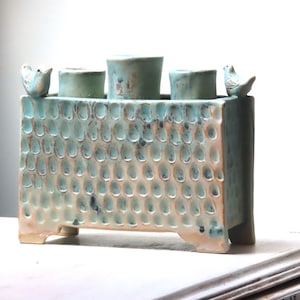 May include: A ceramic candle holder with a textured surface and a turquoise and cream glaze. It has three cylindrical candle holders and two small bird figurines. The piece is handmade and has a unique, artistic design. The dimensions are in centimetres.