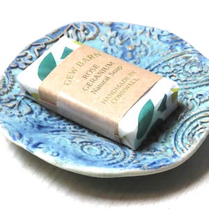 May include: A blue ceramic soap dish with a paisley pattern, featuring a bar of handmade Rose Geranium natural soap wrapped in paper with green leaves.