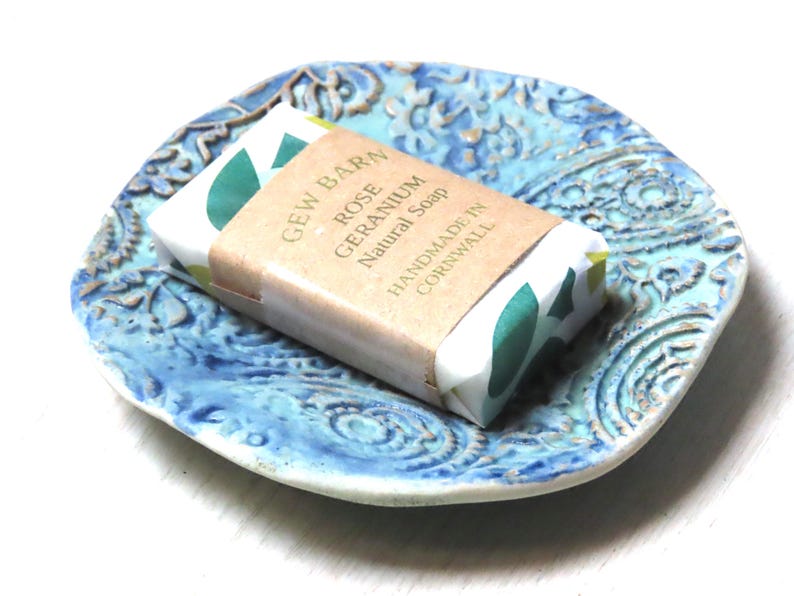 May include: A blue ceramic soap dish with a paisley pattern, featuring a bar of handmade Rose Geranium natural soap wrapped in paper with green leaves.
