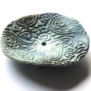 May include: A teal blue ceramic dish with a raised paisley pattern. The dish has a small hole in the center and sits on three small feet.
