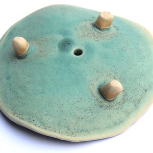 May include: A teal ceramic planter with three small, rounded feet. The planter has a hole in the center for drainage.