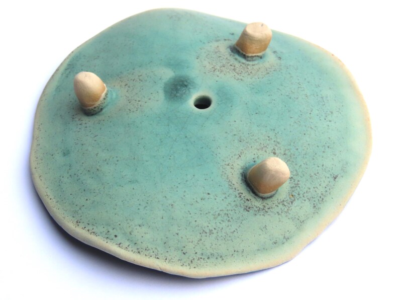 May include: A teal ceramic planter with three small, rounded feet. The planter has a hole in the center for drainage.