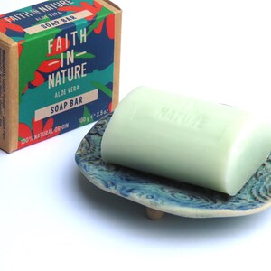 May include: A green soap bar with the words "Faith in Nature" and "Aloe Vera" printed on it. The soap bar is sitting on a blue and white ceramic soap dish.