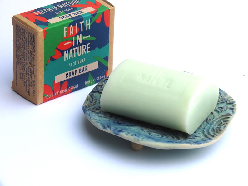 May include: A green soap bar with the words "Faith in Nature" and "Aloe Vera" printed on it. The soap bar is sitting on a blue and white ceramic soap dish.