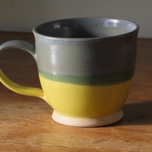 May include: A handmade ceramic mug with a rounded shape and a large handle. The mug features a colour-blocked design with a yellow base, a green stripe, and a grey top. The handle is yellow. The mug sits on a white base.