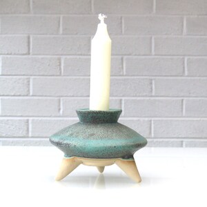 May include: A teal blue ceramic candle holder with three legs and a speckled finish. A white candle sits in the holder.
