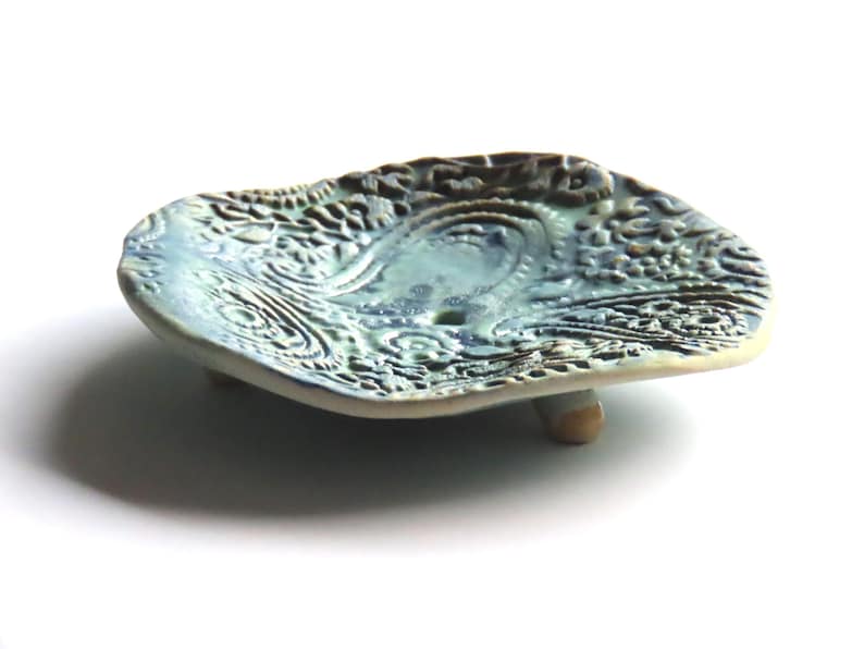 May include: A blue and green ceramic soap dish with a raised, textured design. The dish has three small feet and is shaped like a leaf.